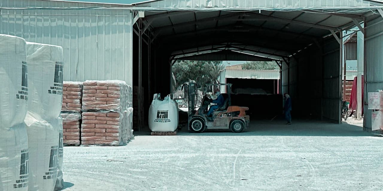 A forklift carrying a large bag labeled 'CMI' drives into a warehouse, with stacks of bags and a worker visible in the background at CMI's Sohar plant.